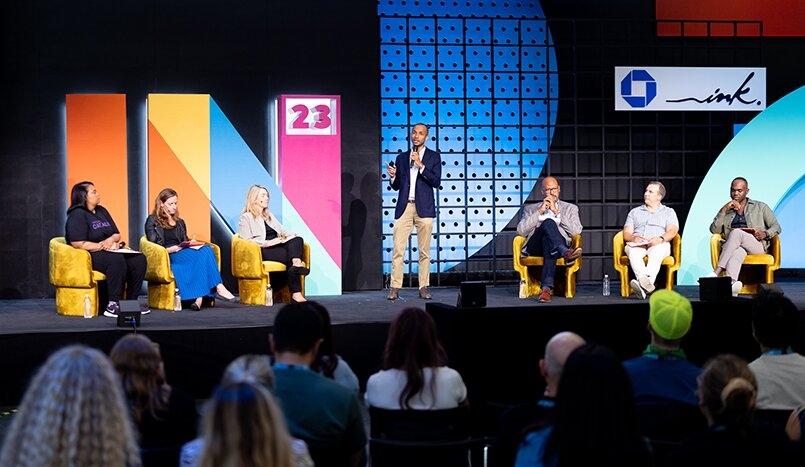 Man talking with six other panelists on a stage in front of an audience