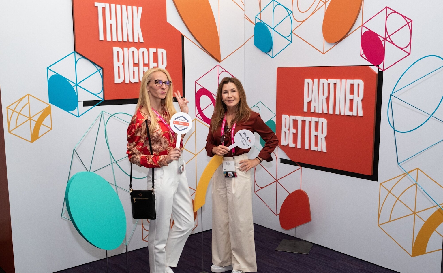 Two people holding signs standing against a backdrop that says "Think Bigger" and "Partner Better".